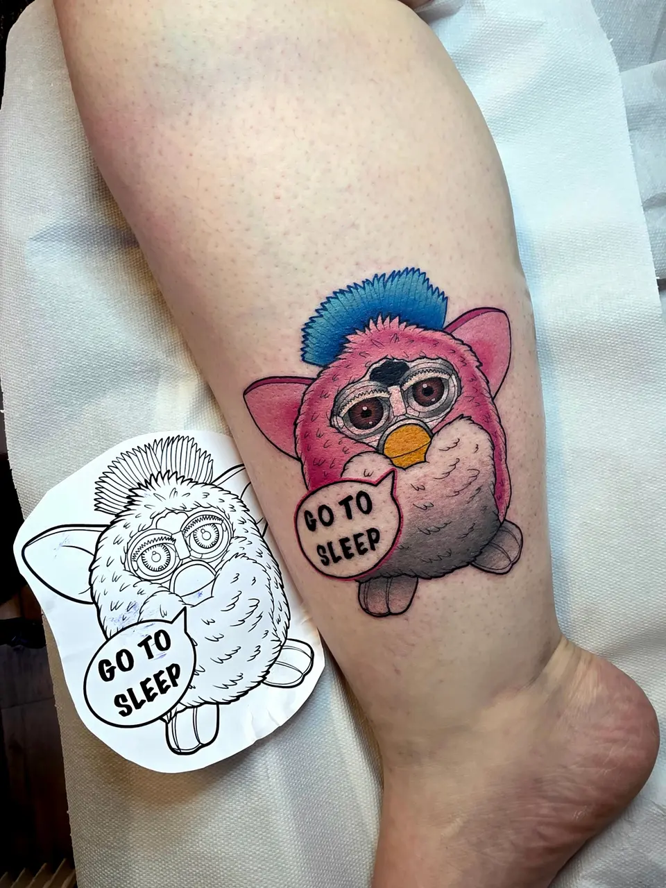 Furby violet « Go to sleep » tatoué sur la jambe, posé à côté de son dessin