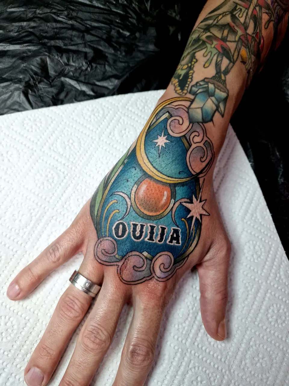 Main tatouée par Ben d'une planchette Ouija bleu canard, avec lune et étoiles