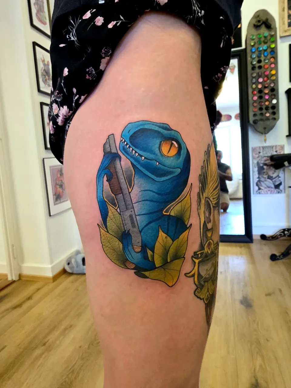 Vélociraptor bleu entouré de feuillage, tatoué sur la cuisse